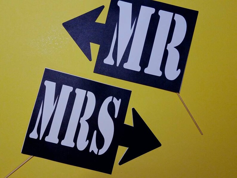 May include: Two black signs with white text that read "MR" and "MRS" with arrow designs. The signs are on wooden sticks and set against a yellow background. Ideal for wedding or engagement photos.