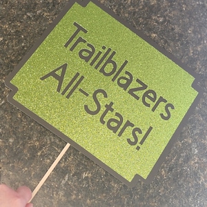 May include: Green glitter cardstock sign with black text that reads "Trailblazers All-Stars!" The sign is attached to a wooden stick.