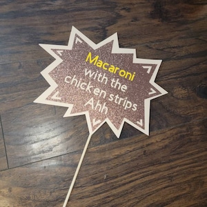 May include: A pink and white star-shaped sign with the text "Macaroni with the chicken strips Ahh" in yellow letters. The sign is on a stick.