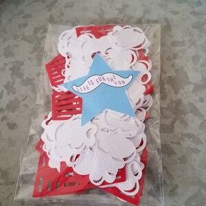 May include: A clear plastic bag filled with red and white paper confetti. The confetti is in the shape of stars, clouds, and rectangles. A blue star with the text "Life is for the fun" and a black mustache is in the center of the bag.