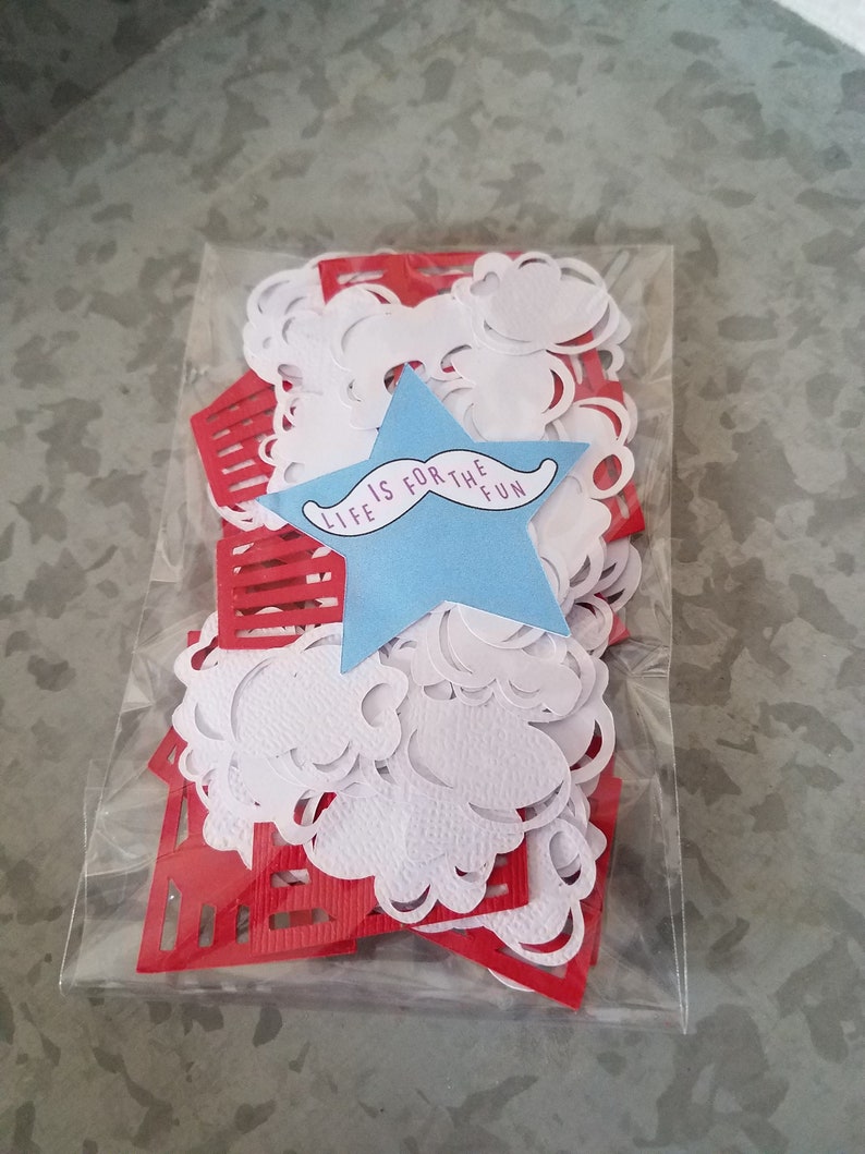 May include: A clear plastic bag filled with red and white paper confetti. The confetti is in the shape of stars, clouds, and rectangles. A blue star with the text "Life is for the fun" and a black mustache is in the center of the bag.