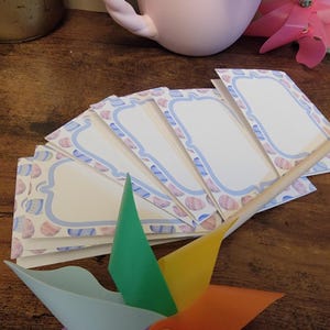 May include: A set of six blank gift tags with a pastel blue and pink seashell design. The tags are arranged in a fan shape and are ready to be written on.