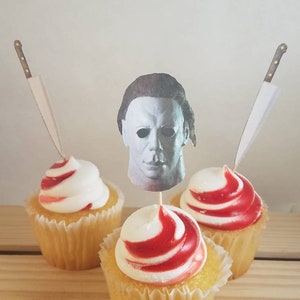 May include: Three cupcakes with red and white swirl frosting. The center cupcake has a paper cutout of Michael Myers' mask from the Halloween movie series.