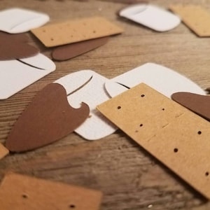 May include: Close-up of scattered paper cutouts resembling s'mores. The cutouts include white marshmallows, brown chocolate pieces, and tan graham crackers with small holes. The items are arranged on a wooden surface.