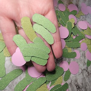 May include: A hand holding a pile of green and gold glitter paper cutouts shaped like pickles. There are also pink glitter paper circles scattered around.