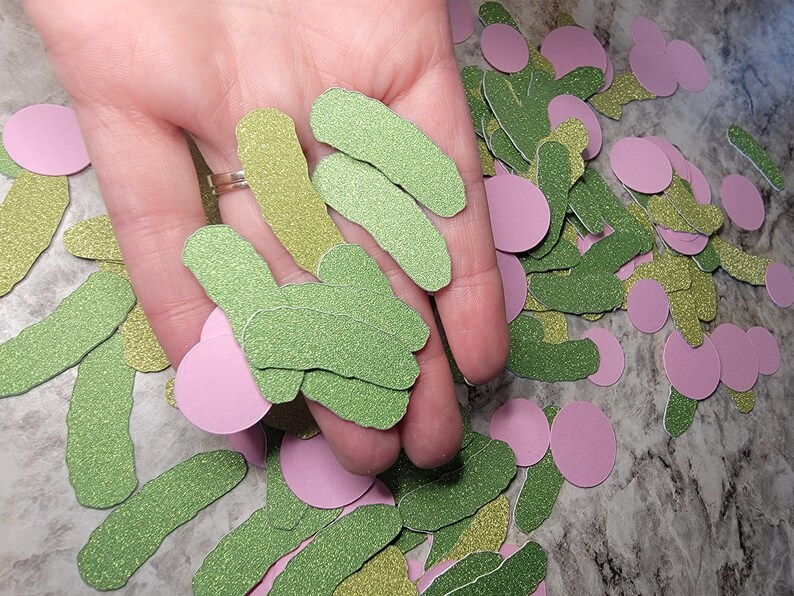 May include: A hand holding a pile of green and gold glitter paper cutouts shaped like pickles. There are also pink glitter paper circles scattered around.