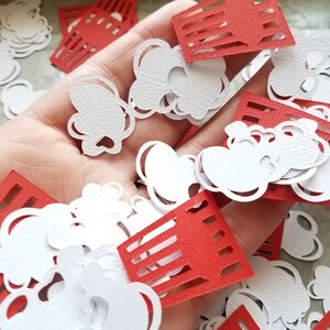 May include: A pile of red and white paper cutouts in the shape of popcorn and popcorn buckets. The cutouts are scattered on a hand.