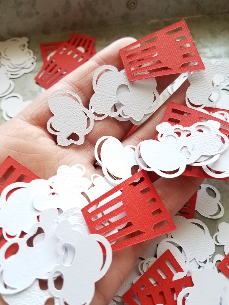May include: A pile of red and white paper cutouts in the shape of popcorn and popcorn buckets. The cutouts are scattered on a hand.
