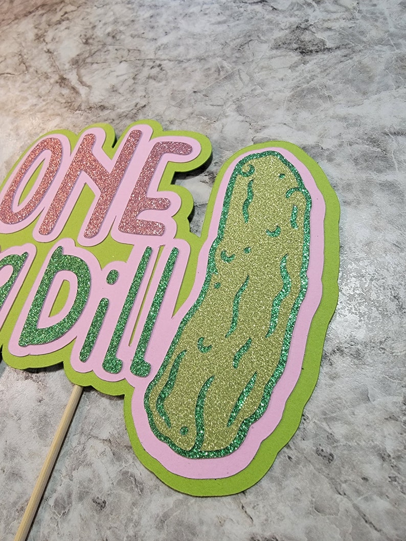 Op de afbeelding: Een groene glitter-augurk met een roze omtrek en de woorden "one dill" in roze glitter op een groene achtergrond.