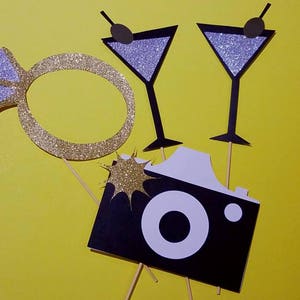 May include: Photo booth props on a yellow background. Includes a gold glitter engagement ring, two martini glasses with glitter, and a black and white camera prop. The martini glasses have black olive accents.