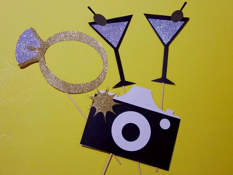 May include: Photo booth props on a yellow background. Includes a gold glitter engagement ring, two martini glasses with glitter, and a black and white camera prop. The martini glasses have black olive accents.