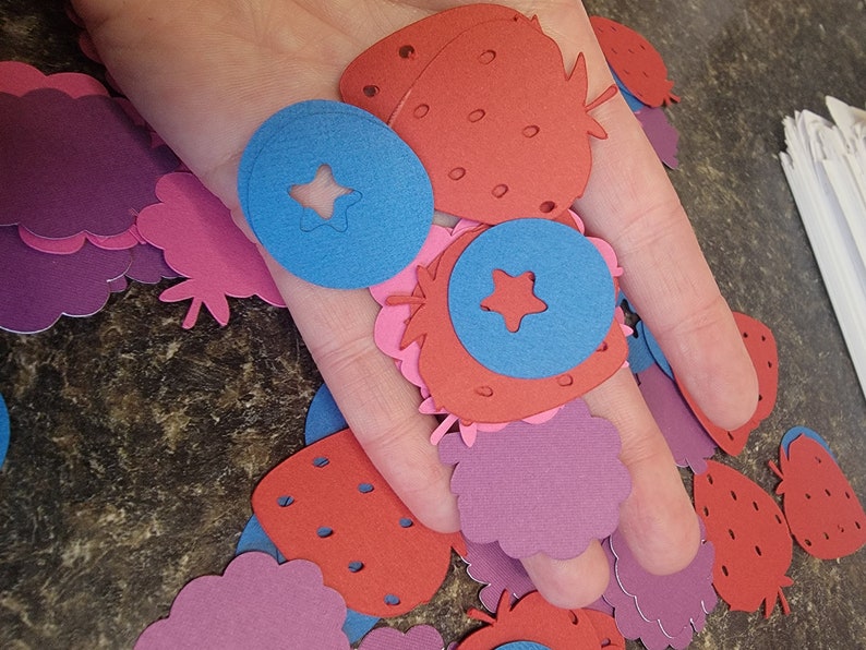 May include: A collection of paper cutouts in the shapes of strawberries, blueberries, and raspberries. The cutouts are in shades of red, pink, and blue.