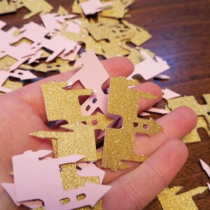 May include: A pile of pink and gold glitter castle confetti. The confetti is shaped like a castle with a pointed roof and a tower.
