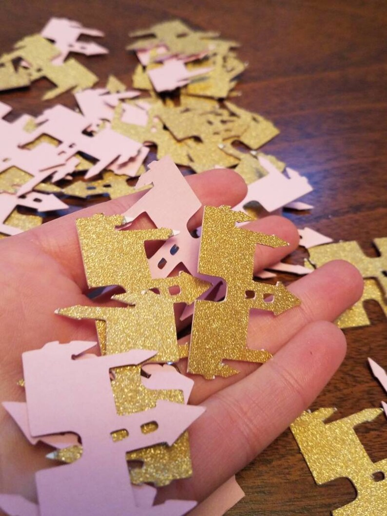 May include: A pile of pink and gold glitter castle confetti. The confetti is shaped like a castle with a pointed roof and a tower.
