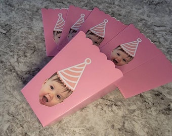 May include: Pink popcorn boxes with a scalloped edge and a cut-out for a photo. Each box has a white and pink striped party hat with a white pom-pom on top. The photo shows a baby wearing a party hat.