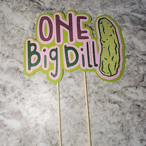 Op de afbeelding: Een groene en roze taarttopper met de tekst "ONE Big Dill" en een illustratie van een groene augurk.