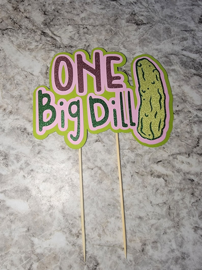 Op de afbeelding: Een groene en roze taarttopper met de tekst "ONE Big Dill" en een illustratie van een groene augurk.