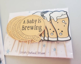 May include: A set of 10 cupcake toppers featuring a beer glass and a speech bubble with the text "A Baby is Brewing". The toppers are on wooden skewers and are packaged in a clear plastic bag.