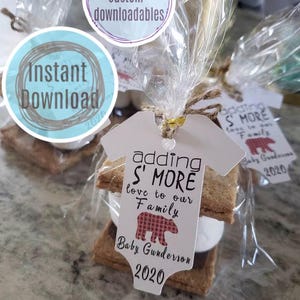 May include: A white tag with a red and black plaid bear design and the text "adding S' MORE love to our Family Baby Gunderson 2020" attached to a s'more treat with graham crackers, marshmallow, and chocolate.