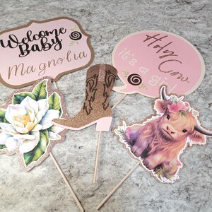 May include: Set of five pink and gold glitter baby shower party decorations. The decorations include a boot, a cow, a flower, and two signs that say "Welcome Baby Magnolia" and "Holy Cow It's a Girl".