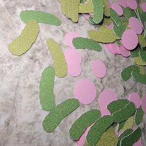 May include: A pile of green and gold glitter pickle shapes and pink glitter circles. These are paper cutouts for a party or craft project.
