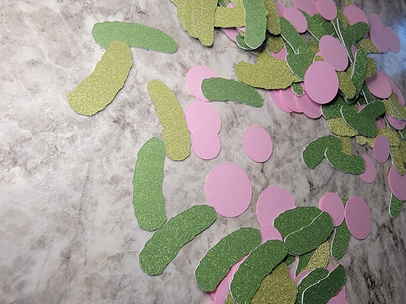 May include: A pile of green and gold glitter pickle shapes and pink glitter circles. These are paper cutouts for a party or craft project.