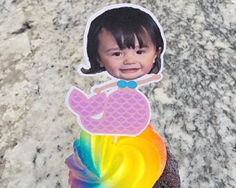 Può includere: Un cupcake al cioccolato con glassa arcobaleno sormontato da un topper per cupcake a forma di sirena con il viso di un bambino.