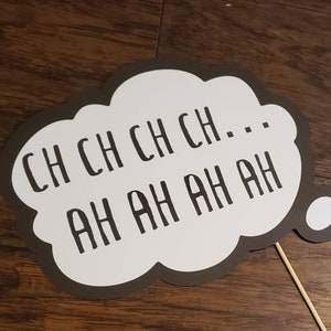 May include: A white speech bubble with a black border and the text "CH CH CH CH... AH AH AH AH" printed in black. The speech bubble is attached to a wooden stick with three white circles.