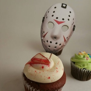 May include: Two cupcakes decorated for a Halloween party. One cupcake has white frosting with red sprinkles and a red candy slice on top. The other cupcake has green frosting with colorful sprinkles. A white hockey mask with black straps and red accents is stuck into the cupcake with white frosting.