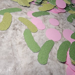 May include: A pile of green and gold glitter pickle shapes and pink glitter circles. The shapes are scattered on a light gray surface.
