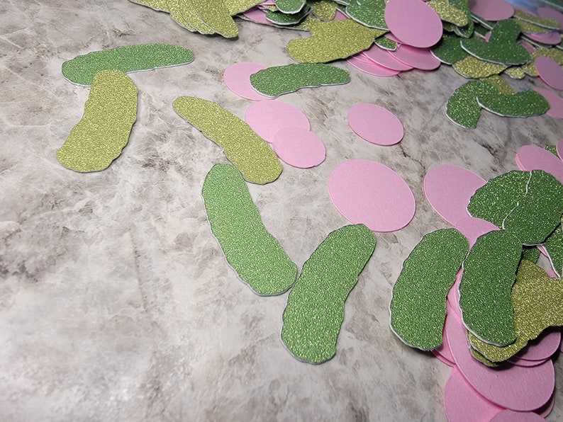 May include: A pile of green and gold glitter pickle shapes and pink glitter circles. The shapes are scattered on a light gray surface.