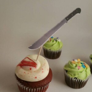 May include: A red velvet cupcake with white frosting and red sprinkles, topped with a fake knife. The cupcake is sitting on a white surface next to two other cupcakes with green frosting.