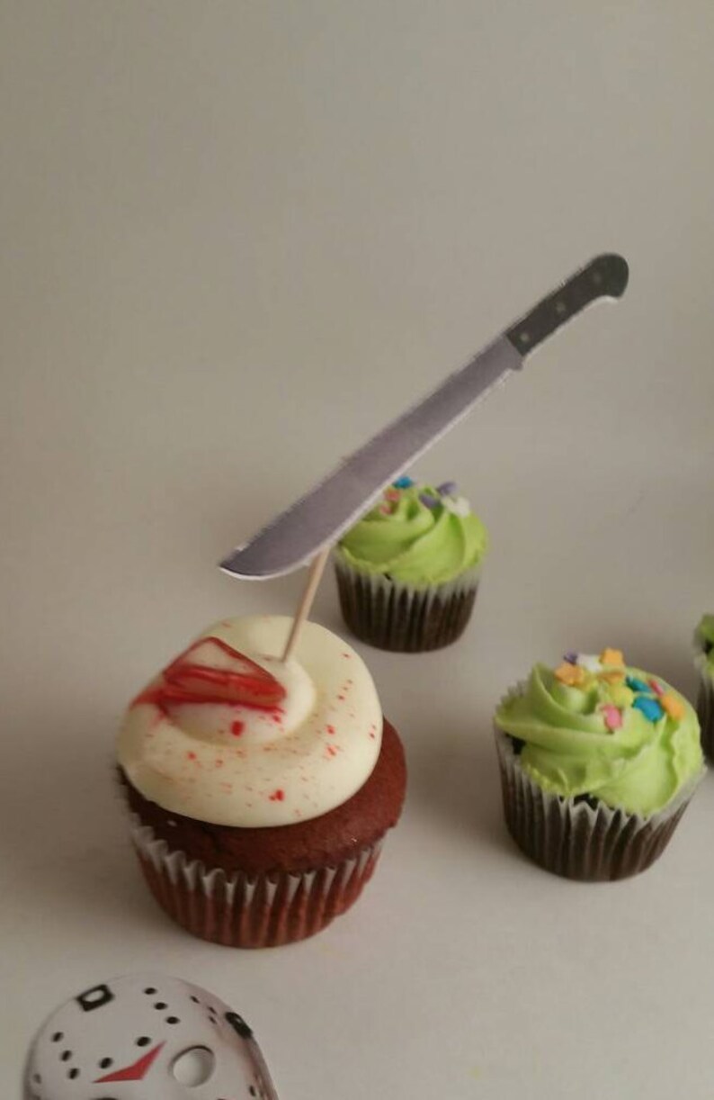 May include: A red velvet cupcake with white frosting and red sprinkles, topped with a fake knife. The cupcake is sitting on a white surface next to two other cupcakes with green frosting.