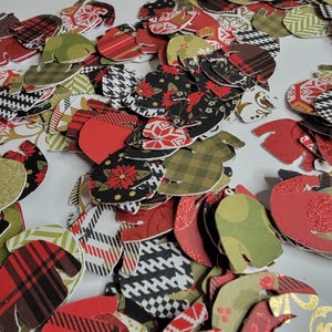 May include: A pile of red, green, and black paper sweater cutouts. The sweaters have different patterns, including plaid, polka dots, and snowflakes.