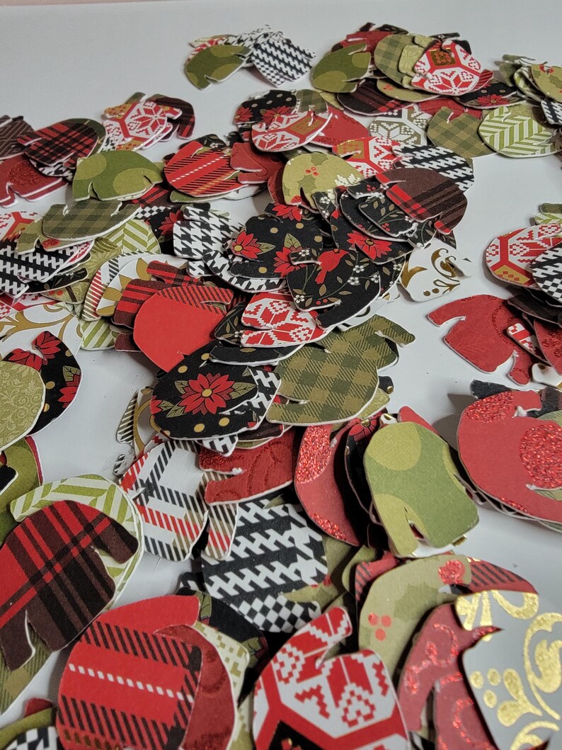 May include: A pile of red, green, and black paper sweater cutouts. The sweaters have different patterns, including plaid, polka dots, and snowflakes.