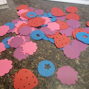 May include: A collection of red, pink, and purple paper cutouts of strawberries, blueberries, and raspberries. The cutouts are scattered on a brown surface.