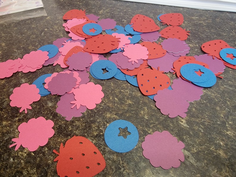 May include: A collection of red, pink, and purple paper cutouts of strawberries, blueberries, and raspberries. The cutouts are scattered on a brown surface.