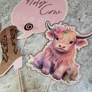 May include: Three pink and gold party decorations for a baby shower. The decorations feature a cow, a boot, and a speech bubble with the text "Holy Cow it's a girl".