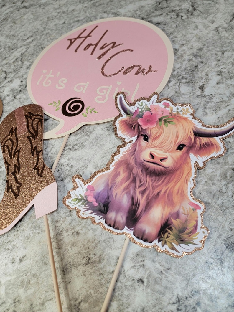 May include: Three pink and gold party decorations for a baby shower. The decorations feature a cow, a boot, and a speech bubble with the text "Holy Cow it's a girl".