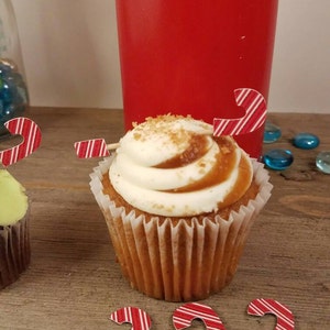 May include: A close-up of a cupcake with white frosting and a caramel drizzle. The cupcake is topped with a red and white candy cane. The cupcake is sitting on a wooden surface with other candy canes.