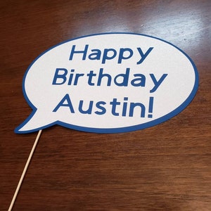 May include: A white speech bubble with a blue border and the text "Happy Birthday Austin!" printed in blue. The speech bubble is attached to a wooden stick.