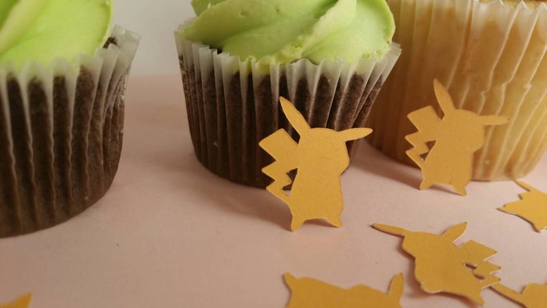 May include: Three cupcakes with green frosting and yellow paper cutouts of a cartoon character on a pink surface.