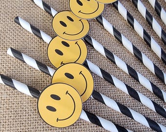 May include: A collection of paper straws with black and white stripes, each adorned with a yellow smiley face. The straws are arranged diagonally on a textured, tan surface, creating a playful and cheerful aesthetic.