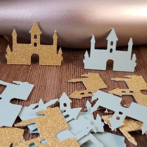 May include: A pile of gold and light blue glitter castle confetti. The confetti is in the shape of a castle with towers and a pointed roof.