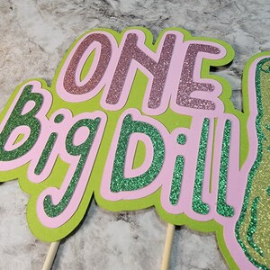 Op de afbeelding: Een groene en roze glittertaarttopper met de woorden "One Big Dill!" erop. De topper zit op twee houten stokjes.