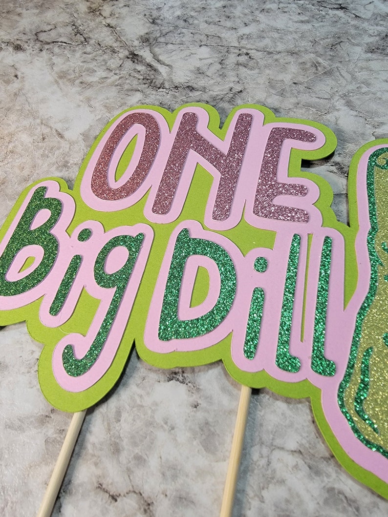 Op de afbeelding: Een groene en roze glittertaarttopper met de woorden "One Big Dill!" erop. De topper zit op twee houten stokjes.
