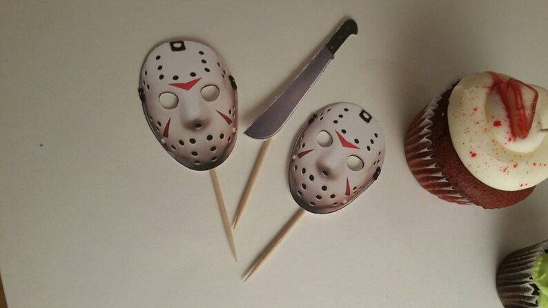 May include: Three cupcake toppers featuring a white hockey mask with red accents, a white hockey mask with red accents, and a silver knife. The toppers are on wooden sticks.