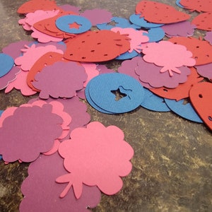 May include: A collection of pink, red, and blue felt cutouts in the shapes of strawberries, flowers, and stars. The cutouts are scattered on a brown surface.