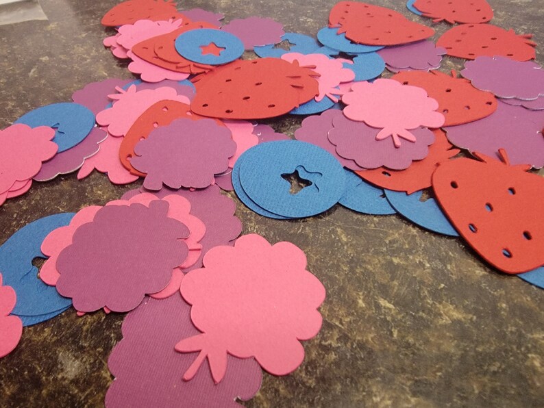 May include: A collection of pink, red, and blue felt cutouts in the shapes of strawberries, flowers, and stars. The cutouts are scattered on a brown surface.