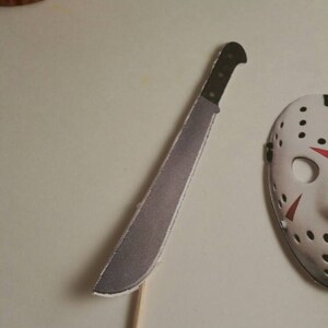 May include: A white cupcake with red frosting and a white cupcake with green frosting. A white paper machete and a white paper hockey mask are on a white surface.